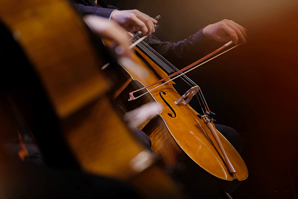 A pair of white hands plays a cello against a dark, softly lit background. Blurred in the foreground, suggesting motion, another pair of hands plays a cello that is much closer in the frame.